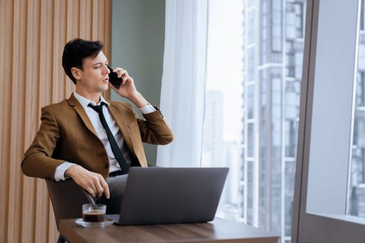 Closeup of handsome businessman making phone call with manager while sitting near window with skyscraper view. Executive manager talking working by using phone and laptop. Look aside. Ornamented.