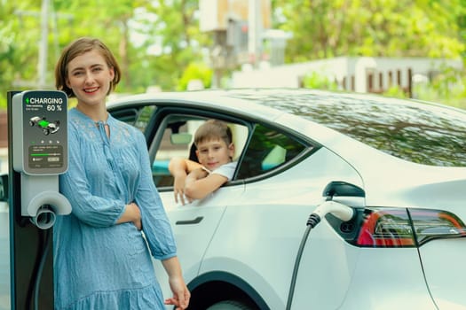 Environmental awareness family with eco-friendly electric car recharging battery from home EV charging station with little boy inside the car. Rechargeable and EV car technology. Perpetual