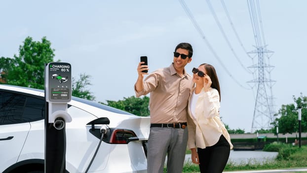 Couple pay for electricity with smartphone while recharge EV car battery at charging station connected to power grid tower electrical as electrical industry for eco friendly car utilization.Expedient