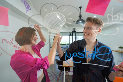 Two smart cooperative business people giving a high five to celebrate the successful start up project while brainstorming marketing idea in front of glass board by using mind map. Immaculate.