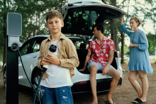 Little boy holding EV charger and point at camera with his family sitting on the trunk in background. Road trip travel with alternative energy charging station for eco-friendly car concept. Perpetual