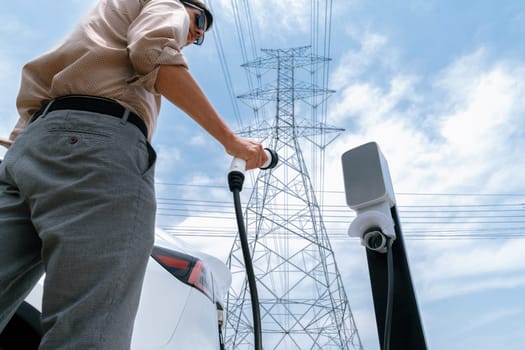 Man recharge EV electric car battery at charging station connected to electrical power grid tower on sky background as electrical industry for eco friendly vehicle utilization. Expedient