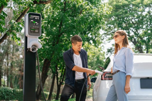 Lovely young couple wearing sun glasses recharging battery for electric car during road trip travel EV car in natural forest or national park. Eco friendly travel during vacation and holiday. Exalt