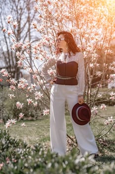 Magnolia park woman. Stylish woman in a hat stands near the magnolia bush in the park. Dressed in white corset pants and posing for the camera