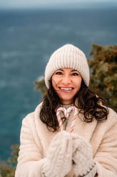 Woman candy sea. Smiling woman in knitted hat, mittens and beige coat holding lollipops candy canes in her hands in shape of heart against the backdrop of the sea