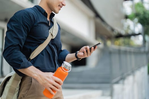 Smiling asian businessman hold reusable eco-friendly ecological cup and using mobile while on the way home at park.