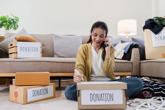 Young woman Asian of charity putting clothes in boxes with donations lettering and using smartphone to contact donations.