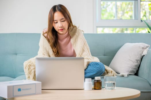 Asian sick woman covered blanket suffering from virus but still working at home with laptop computer, female taking pills for flu while typing laptop keyboard, Unhealthy employee work