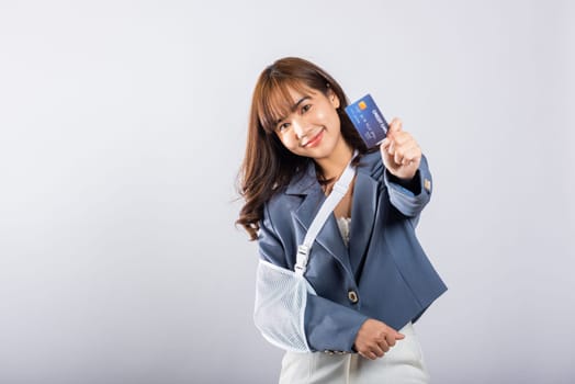 Injured woman smiles through pain of a broken arm, wearing a splint. Emergency medical expenses covered with a credit card. Happy Asian female with sling support isolated on white. Injury is clear.