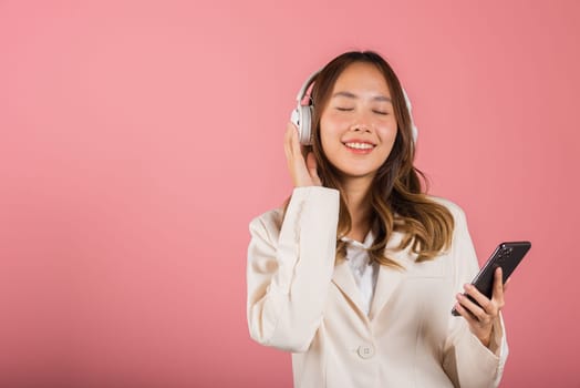 Happy female using mobile phone listens loud song, Portrait of Asian beautiful young woman has fun close eyes listening music with headphones in smartphone app, studio shot isolated on pink background