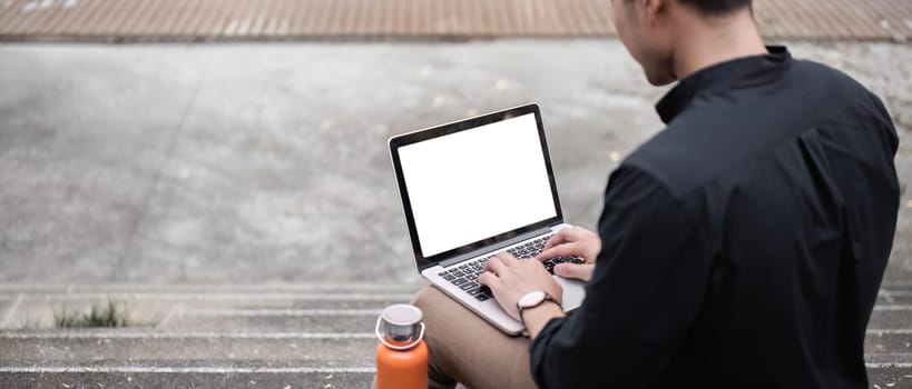 Happy young Asian businessman holding laptop with blank screen outside the office.