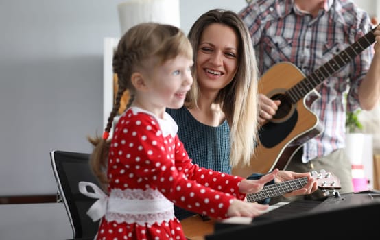 Parents play guitars, daughter on synthesizer. While playing music, child develops mathematical abilities. Child learns to understand music. Parents monitor childs posture during class