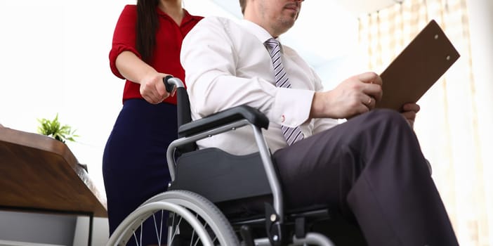 Close-up of woman co-worker accompany man in disabled carriage. Employee reading important papers. Business and adaptation of people with disabilities in society concept