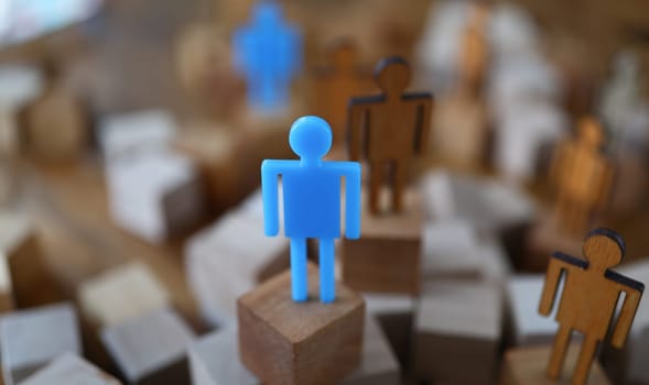 Close-up view of wooden human models placed on blocks. Blue plastic person on centre stage. Stand out from the crowd and leadership and uniqueness concept