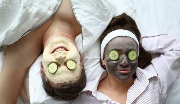 Top view of man and woman in face masks laying on bed. Moisturizing treatment and cucumbers on eyes. Beauty procedure for couple. Skincare and cosmetology concept