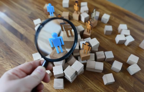 Close-up view of person hand holding magnifying glass and looking through on plastic human model standing on wooden block. Stand out from the crowd concept