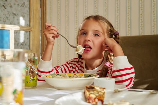 Little cute girl 5 years old with two pigtails eats homemade dumplings with sour cream while sitting at a covered server table in the rays of sunlight in a home interior