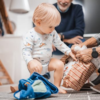 Cute little blond Caucasian toddler unpacking Christmas or Birthday present. Adorable small infant baby boy opening gift.