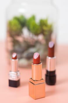 Three pieces of red lipstick stands on a pink and white background with a blur of the decoration of a plant in a jar, side view close-up with depth of field. The concept of cosmetics, beauty salon.