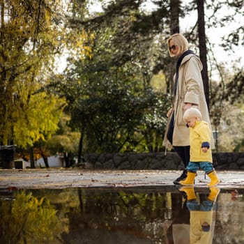 Small bond infant boy wearing yellow rubber boots and yellow waterproof raincoat walking in puddles on a overcast rainy day holding her mother's hand. Mom with small child in rain outdoors