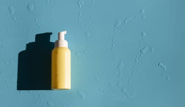 One small yellow bottle with a dispenser and drops of water lies on the left on a blue background with copy space on the right, flat lay close-up. Concept for body care, cosmetics.