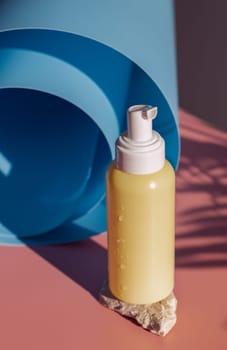 One small yellow bottle with a dispenser, drops of water stands on a stone near a twisted blue roll on a pink background with a palm tree branch, close-up top view. Cosmetics concept, body care.