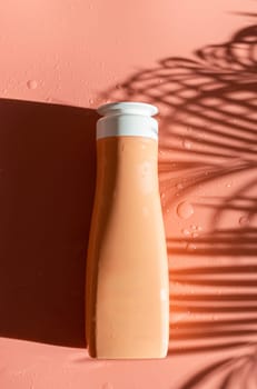 One bottle of liquid soap lies in the center in a plate on a pastel peach background with shadows from a palm branch and drops of water, flat lay close-up.