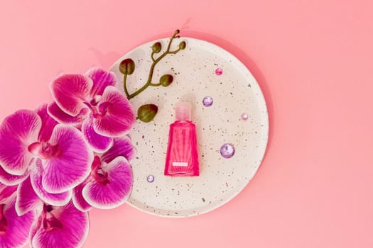 One small pink bottle of liquid soap lies in the center in a plate with one branch of an orchid flower and lilac glass stones on a soft pastel pink background, flat lay close-up.