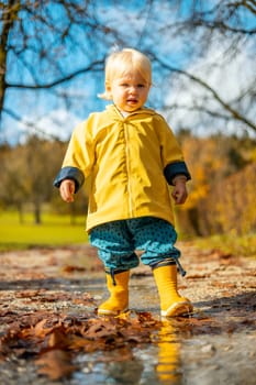 Sun always shines after the rain. Small bond infant boy wearing yellow rubber boots and yellow waterproof raincoat walking in puddles in city park on sunny rainy day