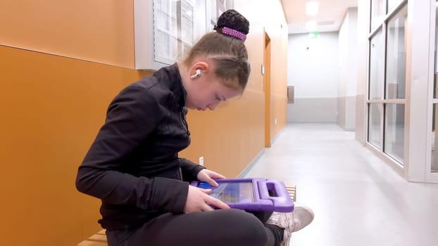 Nestled in a quiet hallway, this dedicated young figure skater finds a moment of tranquility, engrossed in her tablet as she mentally prepares for her upcoming performance.
