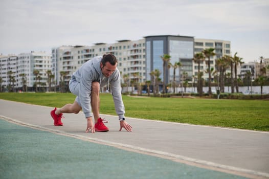 Fit and confident young man runner, standing on the treadmill in starting position ready for running, against urban buildings background. Copy advertising space. Sport. Fitness. Cardio workout