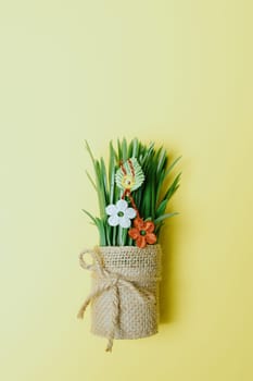 One beautiful homemade martisor of two flowers, a petal and a cheerful smiley face hangs on sprouted wheat in a jute pot lying down on a pastel yellow background with copy space on top, flat lay close-up.