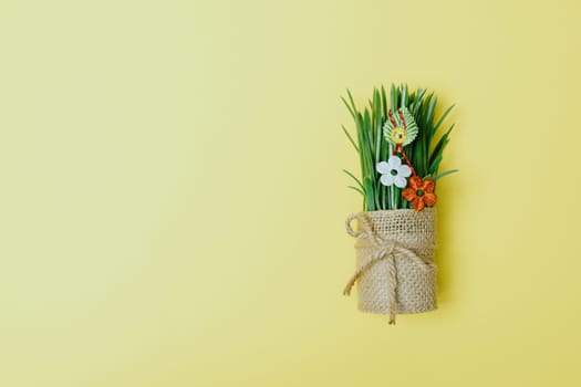 One beautiful homemade martisor of two flowers, a petal and a cheerful smiley face hangs on sprouted wheat in a jute pot lying on the right on a pastel yellow background with copy space on the left, flat lay close-up.