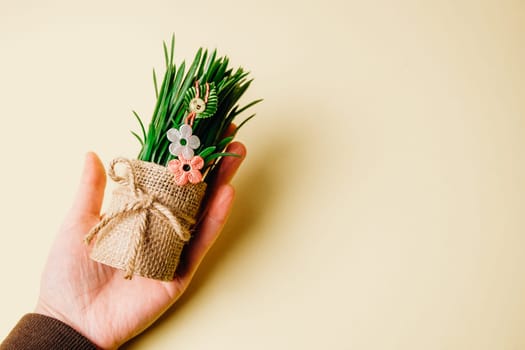 The hand of a young Caucasian woman holds one beautiful homemade martisor of two flowers, a petal and a cheerful smiley face hanging on sprouted wheat in a jute pot on the left on a pastel yellow background with a small copy space sparva, flat lay close-up.
