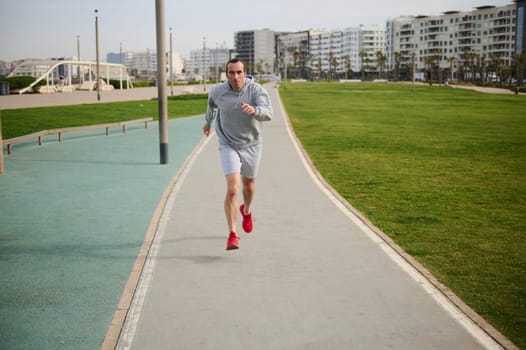 Full length shot of healthy young man in gray sportswear and red sneakers, running on the promenade. Male runner sprinting on the treadmill outdoors.