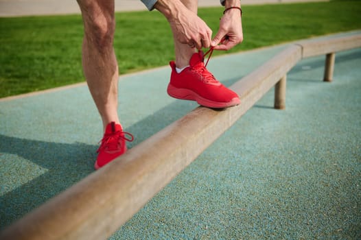 Cropped view of a fit man tying shoelaces of his red sporty sneakers before training or running on the treadmill. Getting ready for morning jog outdoor. Active people and sport concept