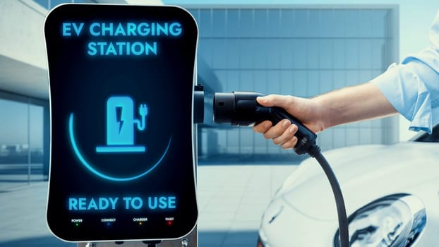 Businessman pull and hold EV charger plug form electric car charging station on sunny modern building in background. Futuristic clean sustainable energy and EV car technological advancement.Peruse