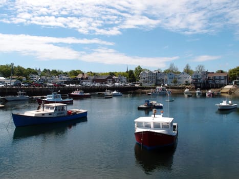 A calm harbor in Rockport, bathed in natural light under a clear blue sky. Boats gently sway in the still waters, their reflections mirroring the peaceful surroundings. The red and blue boats in the foreground add a pop of color against the serene backdrop of buildings and trees. This idyllic harbor invites contemplation and relaxation.
