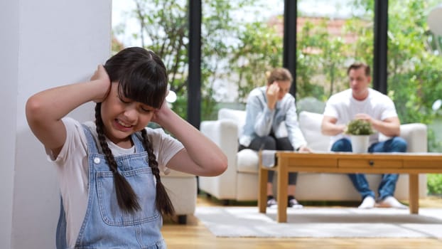 Stressed and unhappy young girl huddle in corner, cover her ears blocking sound of her parent arguing in background. Domestic violence at home and traumatic childhood develop to depression. Synchronos