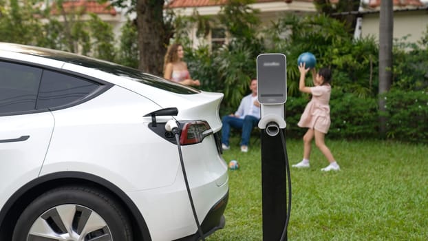 Electric vehicle recharge from home charging station on background of happy and playful family playing together. EV car using alternative and sustainable energy for better future Synchronos