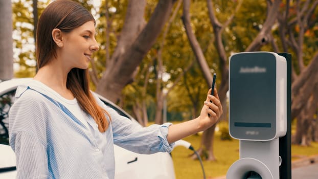 Woman using smartphone online banking application to pay for electric car battery charging from EV charging station during vacation holiday road trip at national park or summer forest. Exalt