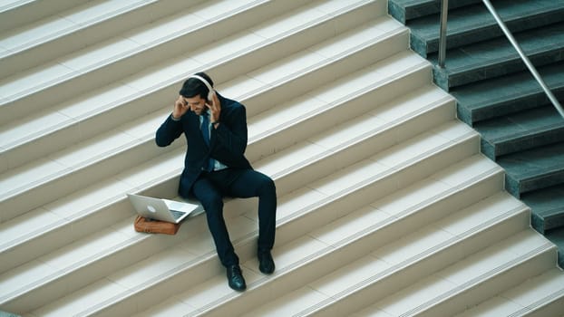 Relaxing smart business man listening music from headphone while sitting at stair. Happy manager smiling while dancing or moving to music. Investor receive good news, getting promotion. Exultant.