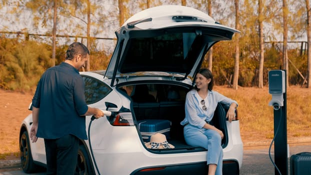 Couple recharge their EV car with electric battery charging station at rest stop during their road trip travel during autumnal season to national park. Relaxing holiday with eco friendly car. Exalt