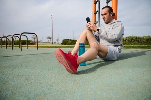 Athletic man checking mobile sports app on his smartphone, analyzing his workout results while sitting on the sportsground after a bodyweight training outdoor. People. Sport and modern technologies