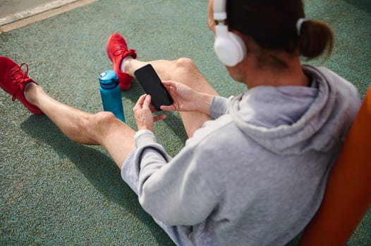 Overhead view of active sports man wearing wireless headphones, checking sports app on his smart mobile phone, sitting on the sportsground. Cell phone with black empty mockup digital touch screen