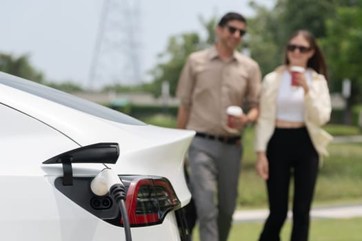 Young couple with coffee cup, recharge electric car's battery from EV charging station in green city park. Sustainable and eco friendly EV car with urban and shopping lifestyle. Expedient