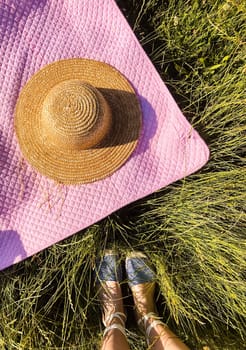 Summer flat lay. Picnic blanket, hat, woman legs on green grass. Top view from above. Summer holiday vacation resting concept. Happy sunshine day. straw hat lies on pink blanket. Girl shoes sandals