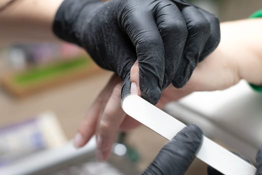 Beauty concept. A manicurist in black latex gloves makes a hygienic manicure, paints the client's nails with gel polish and files them with a nail file in a beauty salon. Close-up. Horizontal.