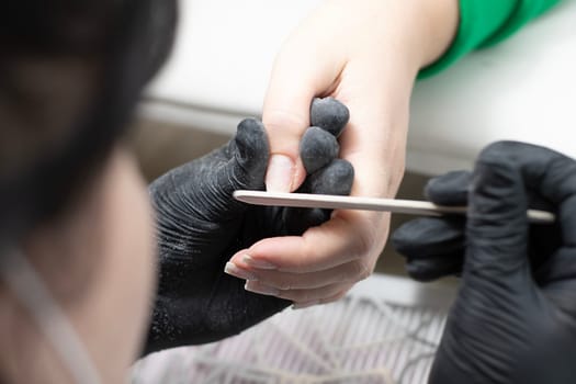 Beauty concept. A manicurist in black latex gloves makes a hygienic manicure, paints the client's nails with gel polish and files them with a nail file in a beauty salon. Close-up. Horizontal.