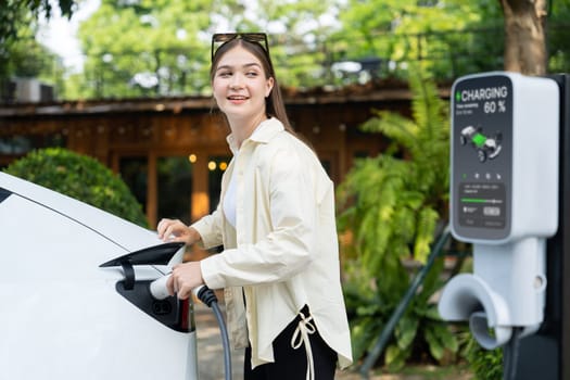 Young woman and sustainable urban commute with EV electric car recharging at outdoor cafe in springtime garden, green city sustainability and environmental friendly EV car. Expedient